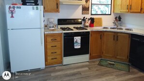 A kitchen with wooden cabinets, a refrigerator, a stove, and a window with a view of trees. - Cozy Milbridge Country Home (Milbridge)