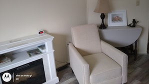 A living room with a white entertainment unit, a beige armchair, a lamp, and a window with a view of greenery. - Cozy Milbridge Country Home (Milbridge)