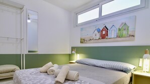 Una camera d'albergo con un letto, un quadro raffigurante delle casette sulla spiaggia, una finestra e un comodino con una lampada.