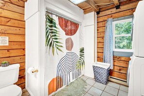 Un baño con detalles de madera, un inodoro blanco, una cortina de ducha con estampado de hojas, una ventana con vistas a árboles y un cubo de basura.