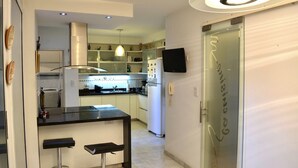 A modern kitchen with a central island, a dining area with bar stools, a built-in oven, and a flat-screen TV mounted on the wall.