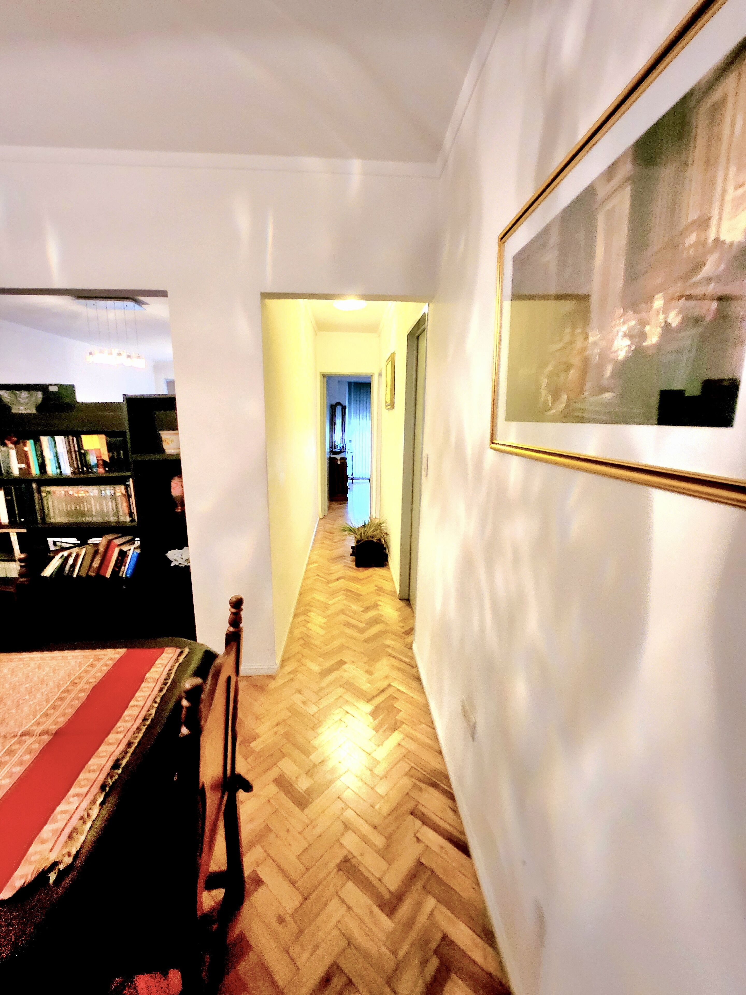 Ein Flur mit Holzdielen im Fischgrätmuster, einem Bett mit roter und grüner Tagesdecke, einem Bücherregal und einem gerahmten Bild an der Wand.