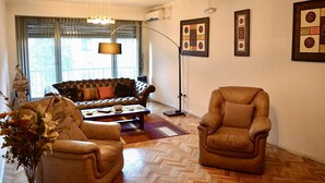 A living room with a sofa, two armchairs, a floor lamp, and a coffee table. There are framed pictures on the wall and a vase with flowers on the floor.
