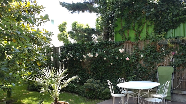 An outdoor patio with a round table and four chairs, surrounded by lush greenery and flowering plants.