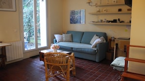 A living room with a sofa, a dining table with chairs, a window with a view of greenery, and shelves with various items.