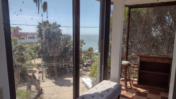 Ein Zimmer mit großem Fenster und Blick auf das Meer und Palmen. Eine weiße gepolsterte Bank und ein Holzsessel sind vorhanden.