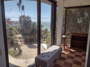 Ein Zimmer mit großem Fenster und Blick auf das Meer und Palmen. Eine weiße gepolsterte Bank und ein Holzsessel sind vorhanden.