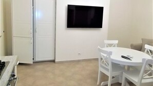Een compacte kamer met een witte eettafel, witte stoelen, een flatscreen televisie aan de muur en een witte deur.