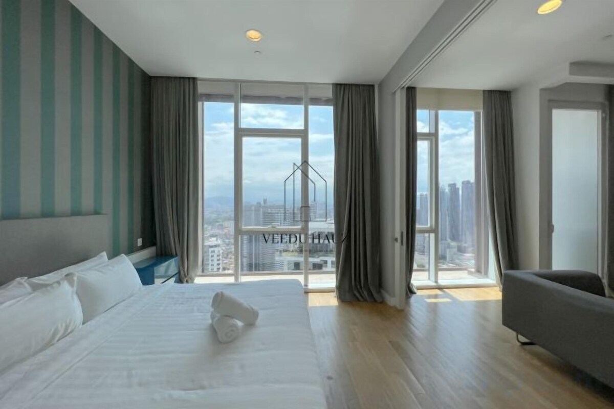 A hotel room with a large bed, a sofa, and a view of a cityscape through large windows.
