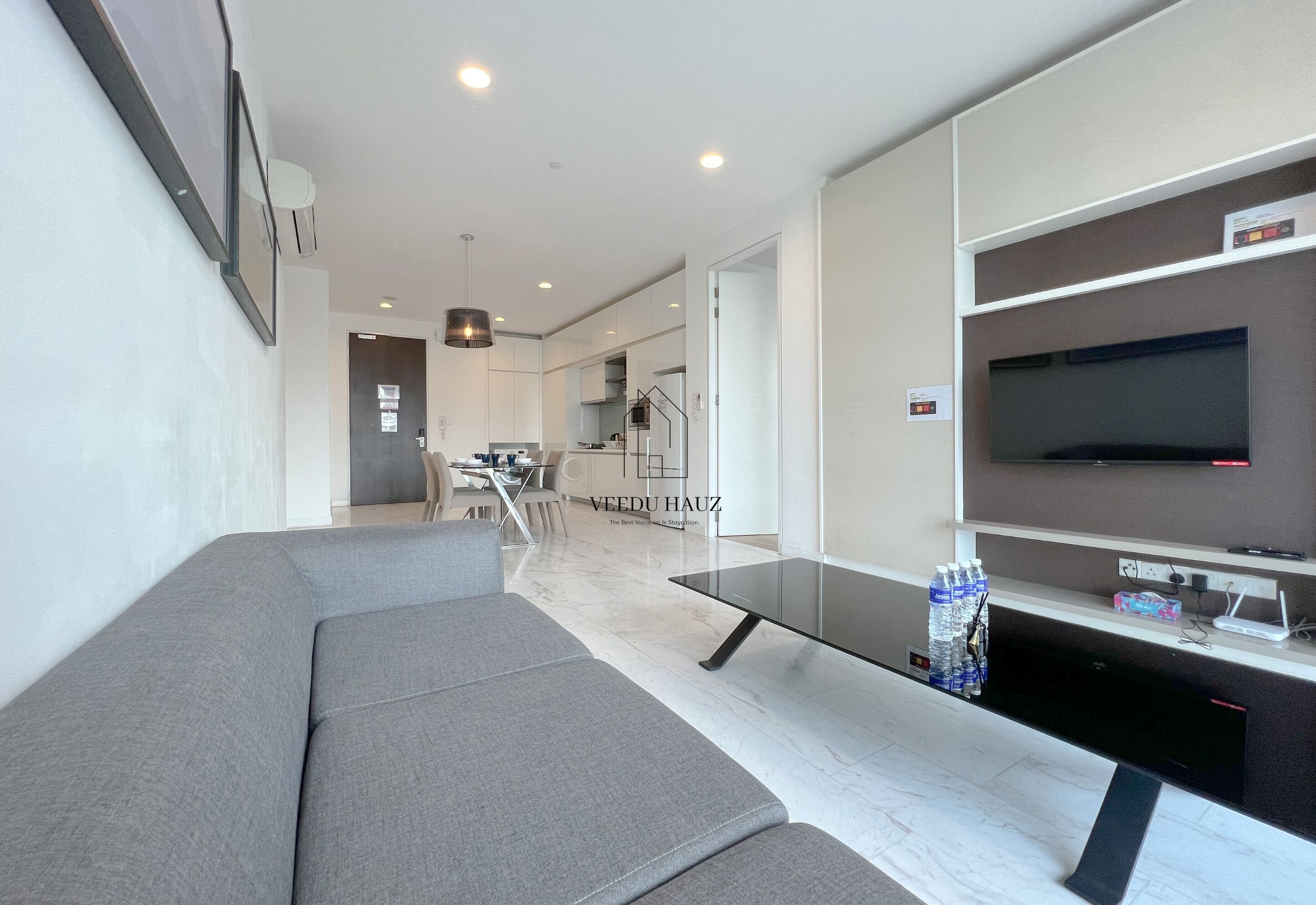 A modern living room with a grey sofa, a glass coffee table, and a flat-screen TV mounted on the wall.