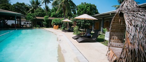 Un'area piscina con poltrone a sdraio e ombrelloni, circondata da palme e una capanna con tetto di paglia.