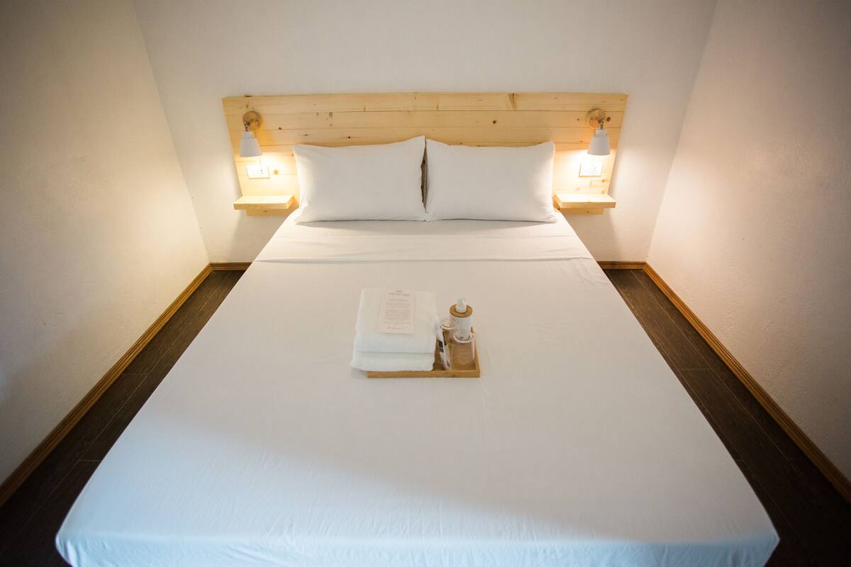 A neatly made bed with white linens, a wooden headboard, and two bedside lamps. A tray with a book and a bottle is placed on the bed.