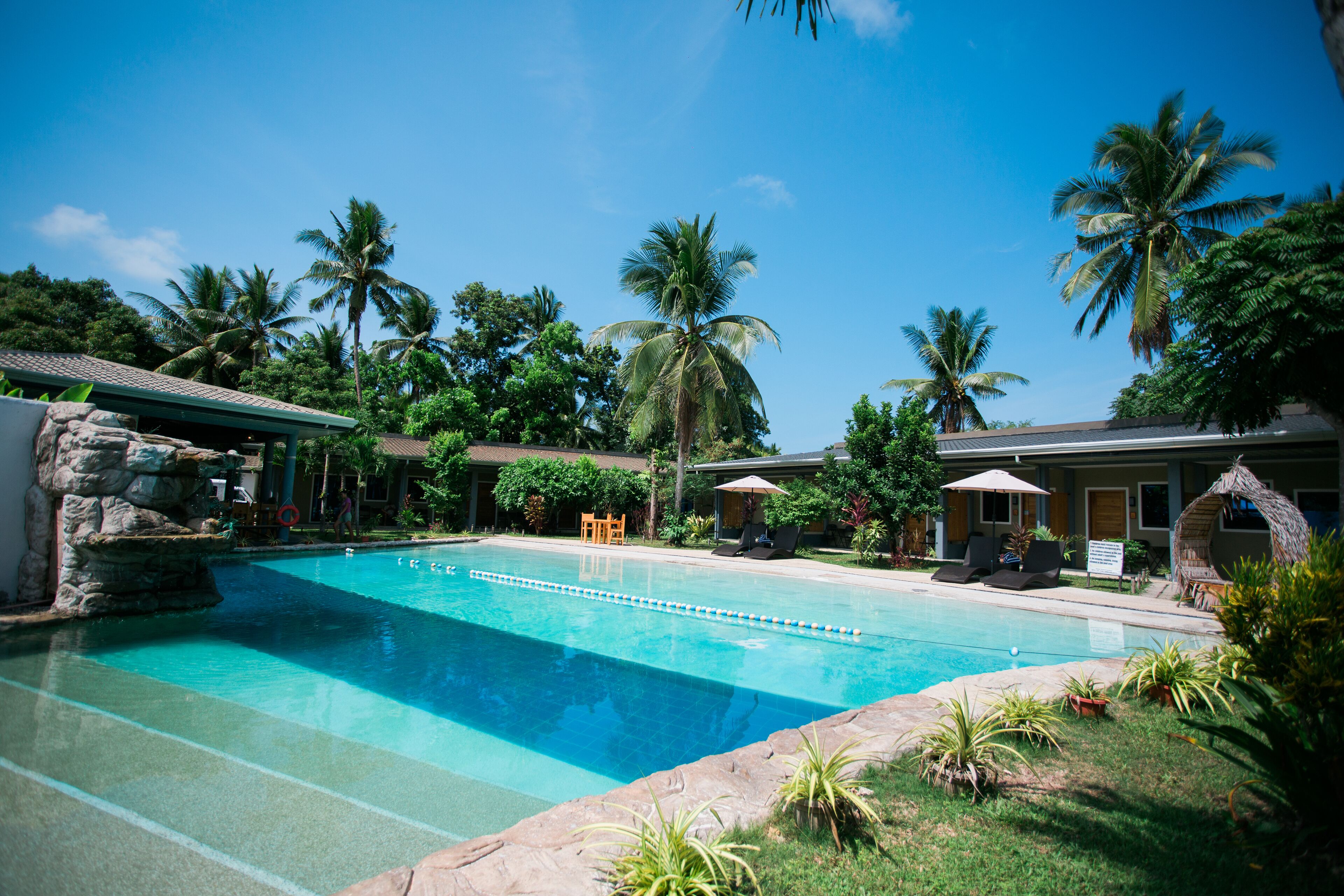 A clear blue pool with lounge chairs and umbrellas, surrounded by palm trees and a stone structure.