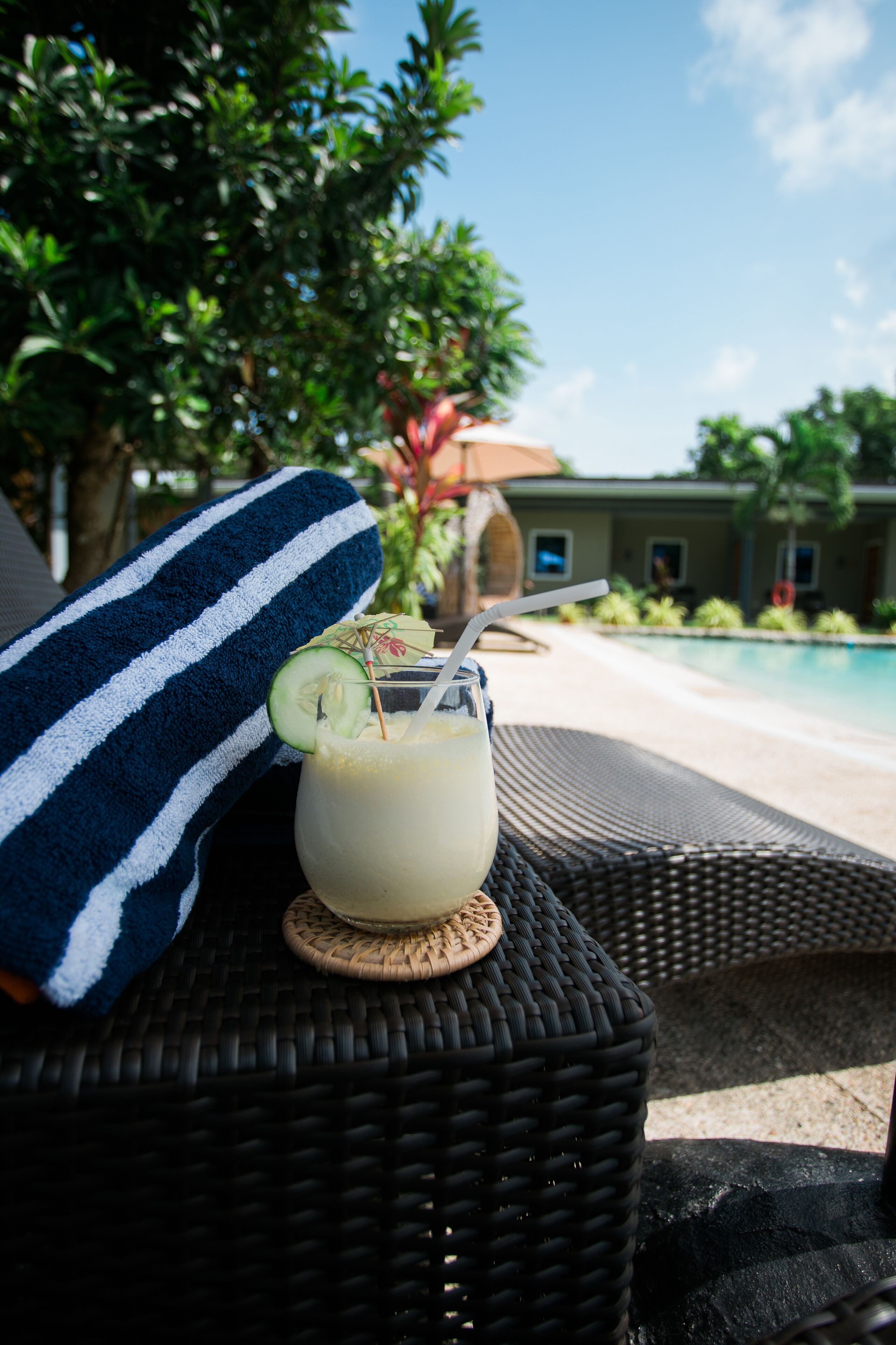 Ein Korbstuhl mit einem Handtuch, ein Getränk mit Trinkhalm und eine Gurkenscheibe, im Hintergrund ein Pool.