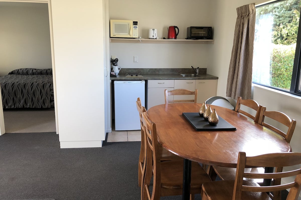 Eine kompakte Küche mit Mikrowelle, Toaster und Wasserkocher auf einem Regal, einem kleinen Kühlschrank und einem Essbereich mit Tisch und Stühlen.