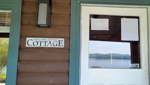 Een houten cottage met een bord waarop 'WELCOME TO THE COTTAGE' staat, een raam met uitzicht op een meer en heuvels, en een politiemelding op het raam.