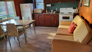 Een keuken met houten kastjes, een koelkast, een magnetron en een fornuis. Er is een eettafel met stoelen en een bank met een kussen.