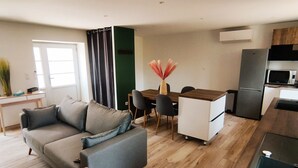 Una sala de estar moderna con un sofá gris, una mesa de comedor con sillas, un área de cocina con refrigerador y microondas, y piso de madera.