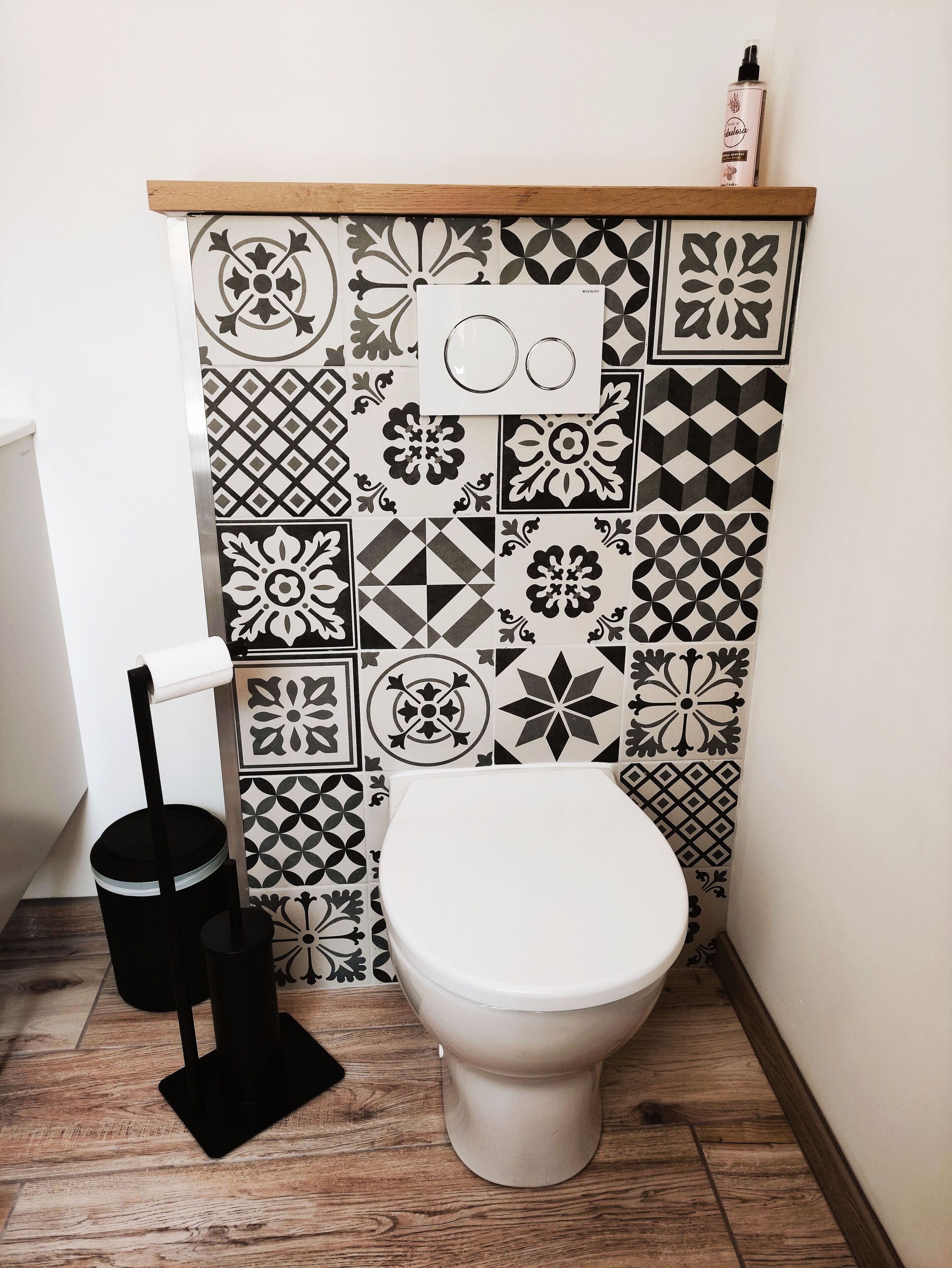 Une salle de bain avec des toilettes, une poubelle noire et un rouleau de papier toilette. Le mur est décoré de différents carreaux noirs et blancs.