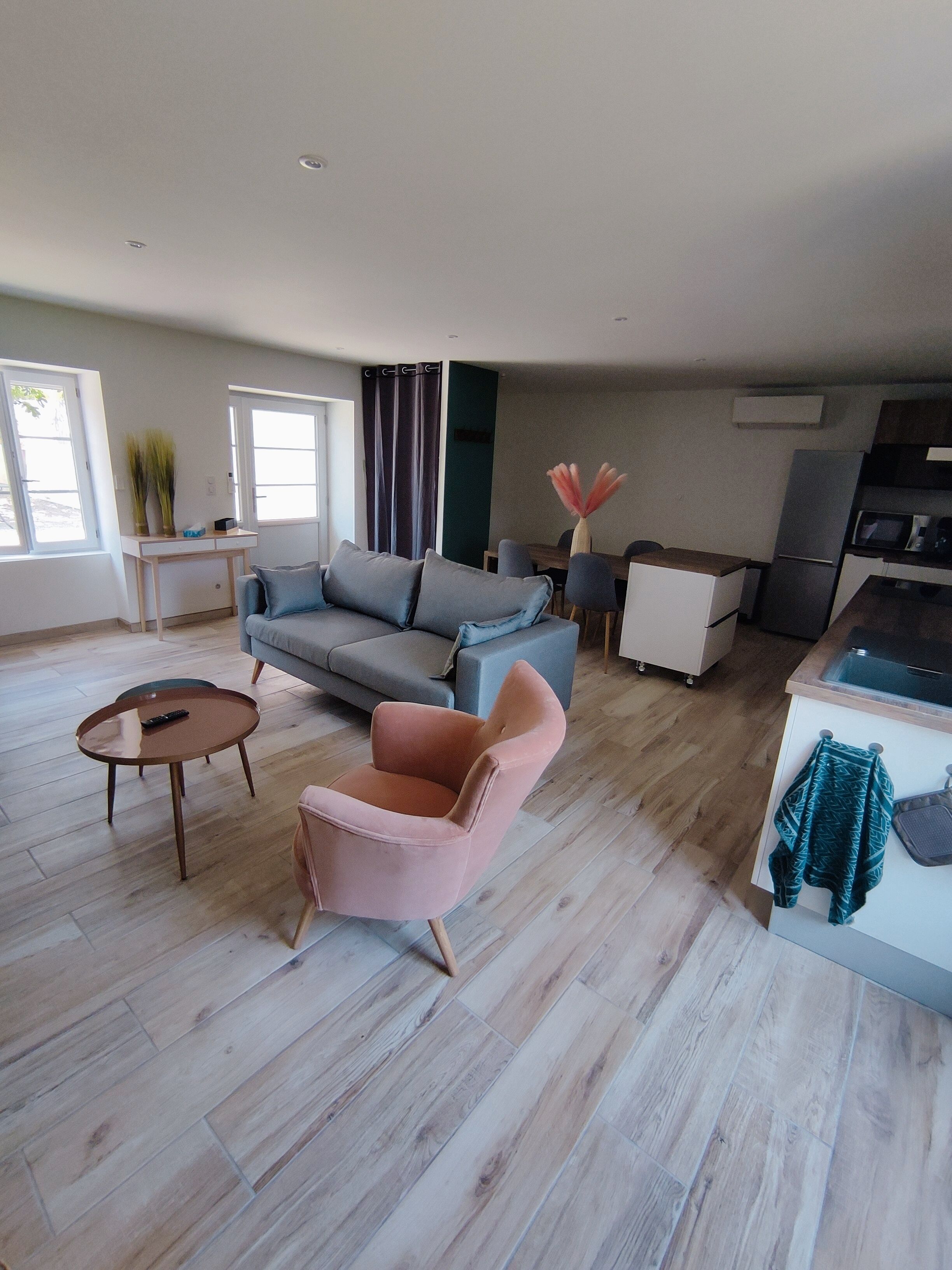 Een moderne woonkamer met een grijze bank, een roze fauteuil en een salontafel. Er is een zithoek met een tafel en stoelen, en een keuken met een koelkast en magnetron.