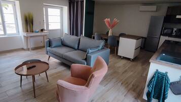 Een moderne woonkamer met een grijze bank, een roze fauteuil en een salontafel. Er is een zithoek met een tafel en stoelen, en een keuken met een koelkast en magnetron.