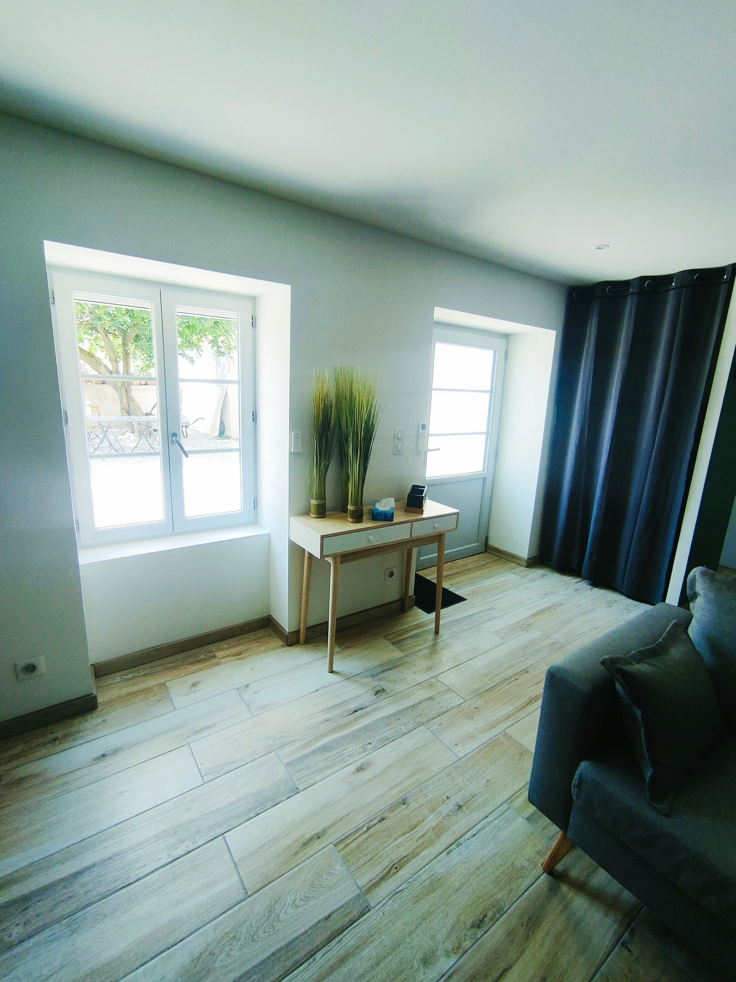 Un salón amplio con suelo de madera, un sofá gris y dos ventanas con cortinas.