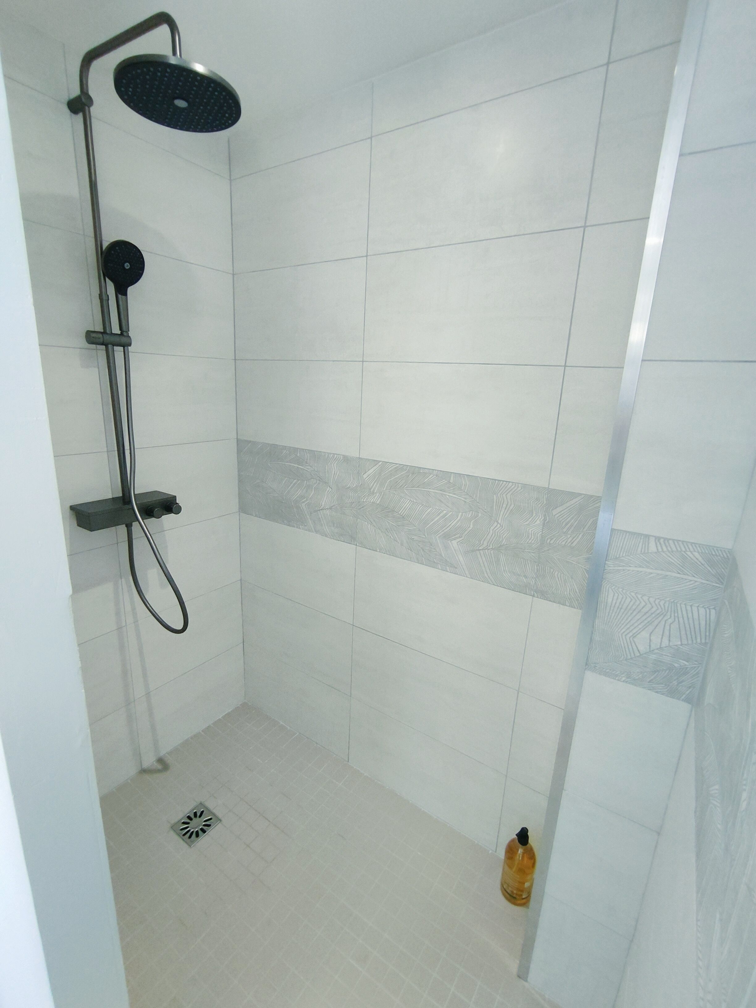 A shower area with a handheld showerhead, a wall-mounted control panel, and a floor drain.