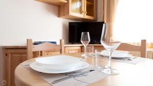Una mesa de comedor dispuesta con platos, vasos y cubiertos, con un televisor y armarios de madera al fondo.
