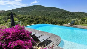 En swimmingpool med en træterrasse, omgivet af livlige lyserøde blomster og et bjerg i baggrunden.