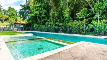 A rectangular swimming pool with clear blue water, surrounded by a paved deck and a metal fence.