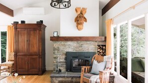 A cozy living room with a stone fireplace, wooden furniture, and a large window with a view of greenery.