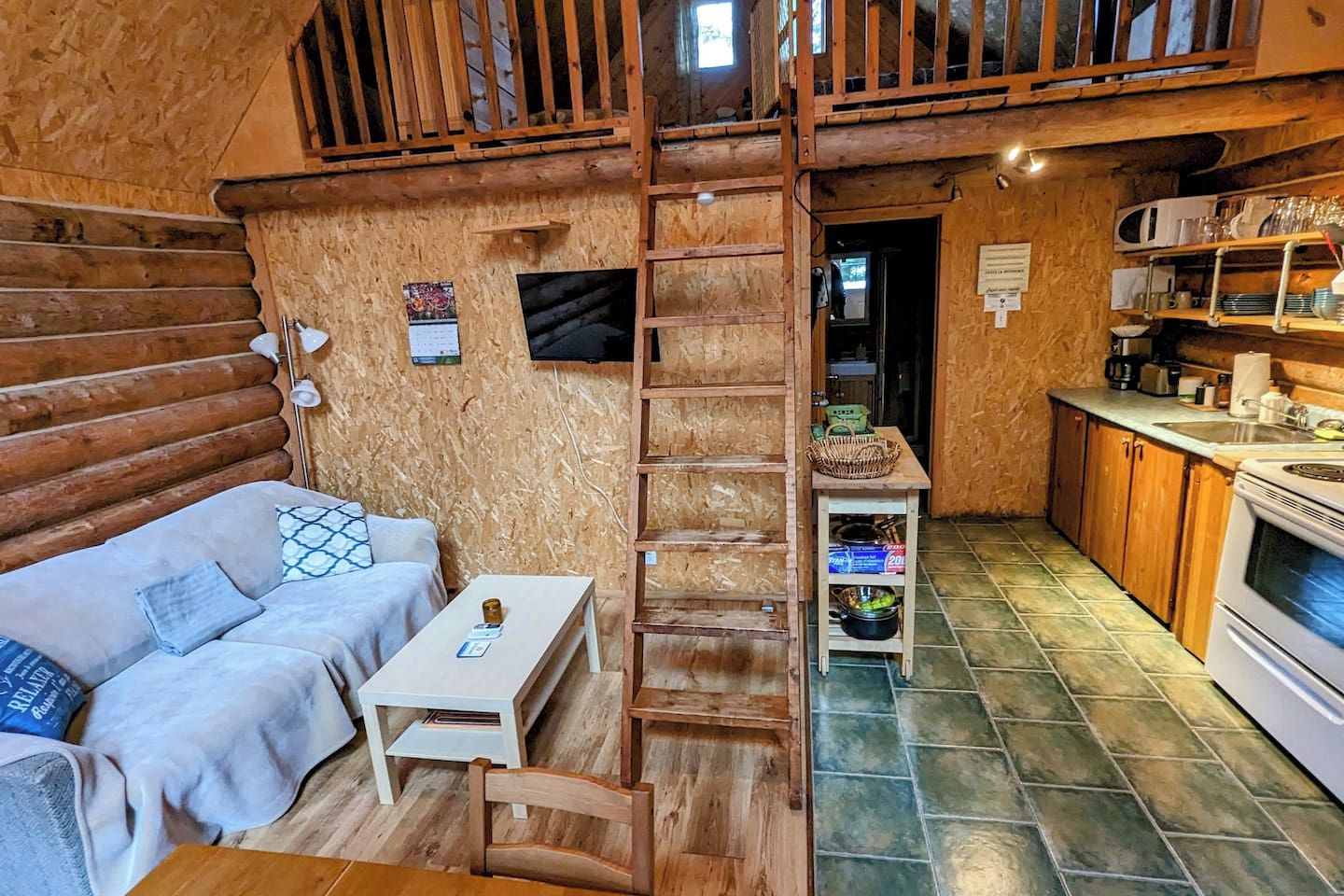 Interior acogedor de una cabaña de troncos con un dormitorio en el altillo, una kitchenette y una sala de estar con sofá y mesa.
