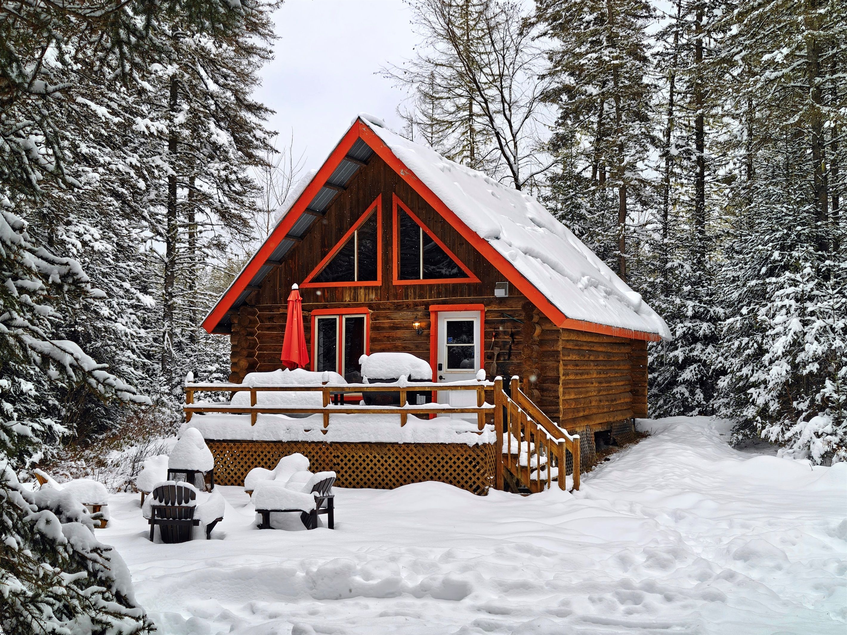 Una cabaña de troncos con un porche cubierto de nieve y un techo rojo, rodeada de un bosque.