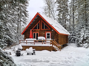 A log cabin with a snow-covered porch and a red roof, surrounded by a forest.