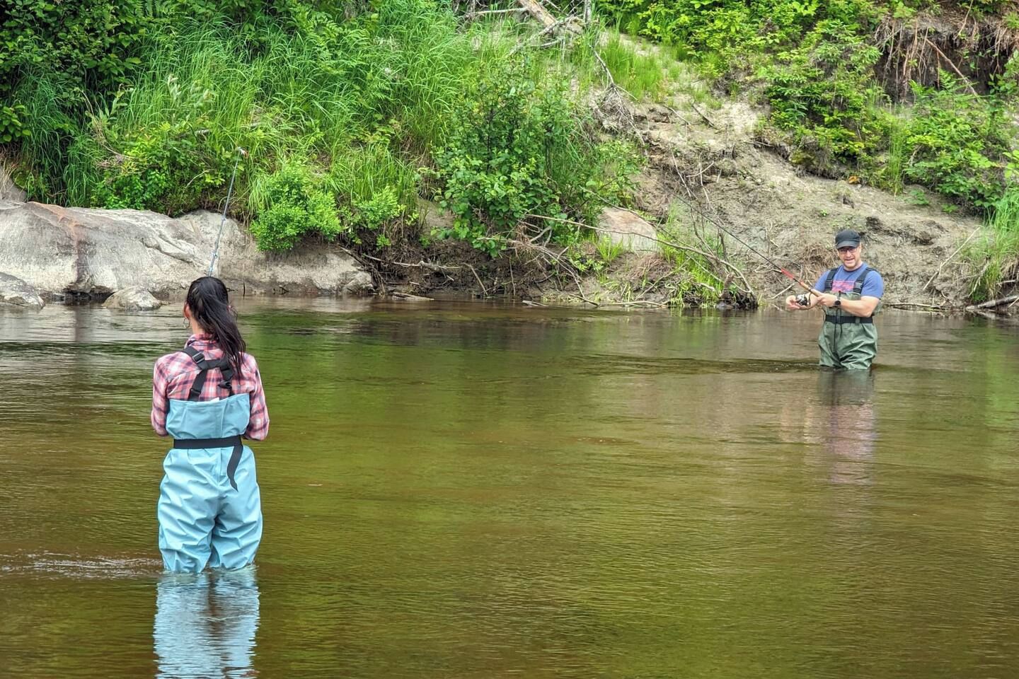 Two individuals fishing in a river surrounded by greenery.