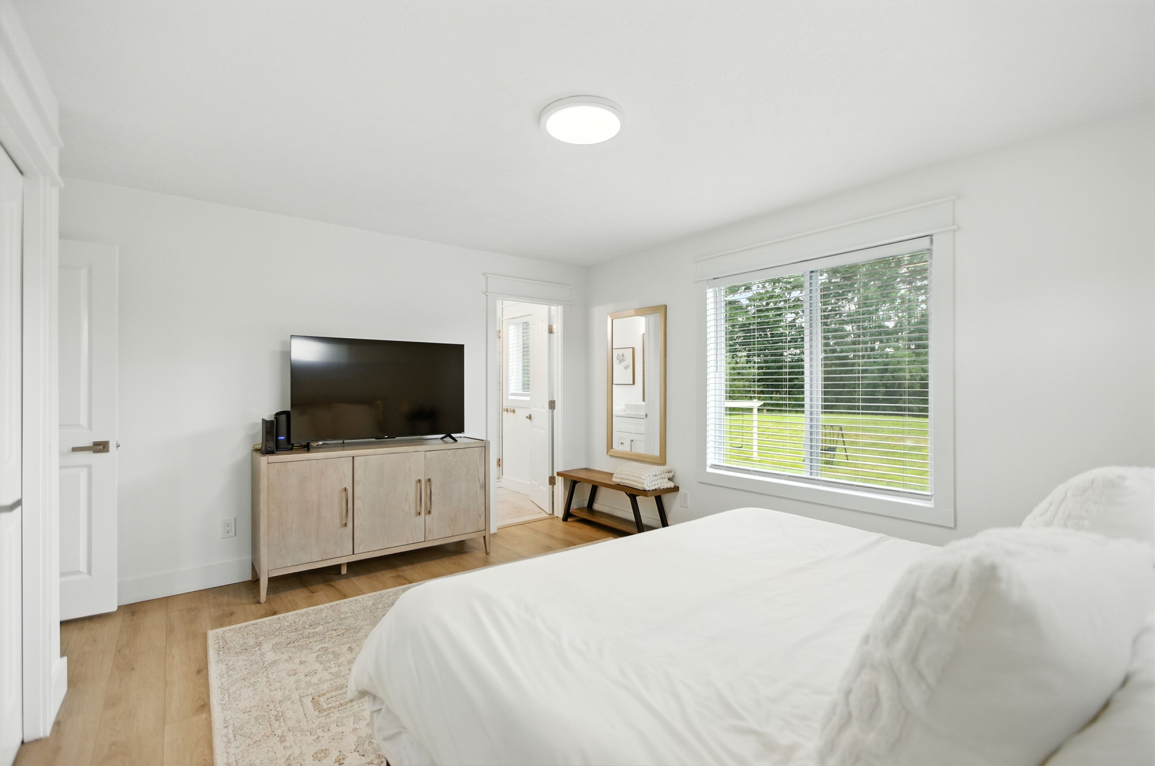 Une chambre à coucher avec un lit, un téléviseur posé sur un meuble en bois, un miroir et une fenêtre avec des stores.