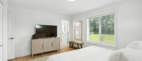 Une chambre à coucher avec un lit, un téléviseur posé sur un meuble en bois, un miroir et une fenêtre avec des stores.