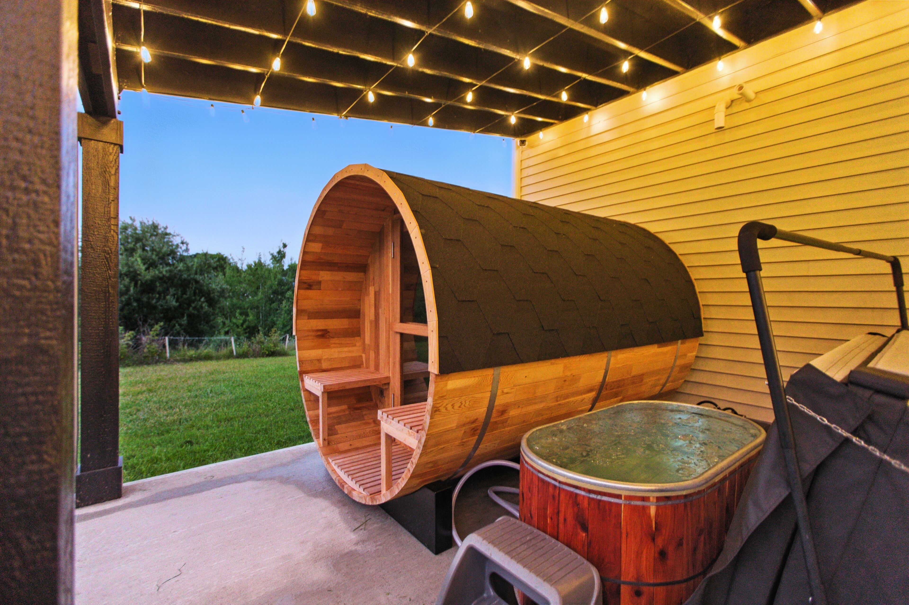 A wooden barrel structure with a bench inside, a hot tub, and a pergola with string lights.