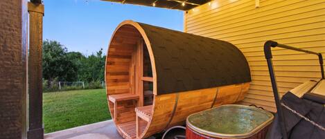 A wooden barrel structure with a bench inside, a hot tub, and a pergola with string lights.