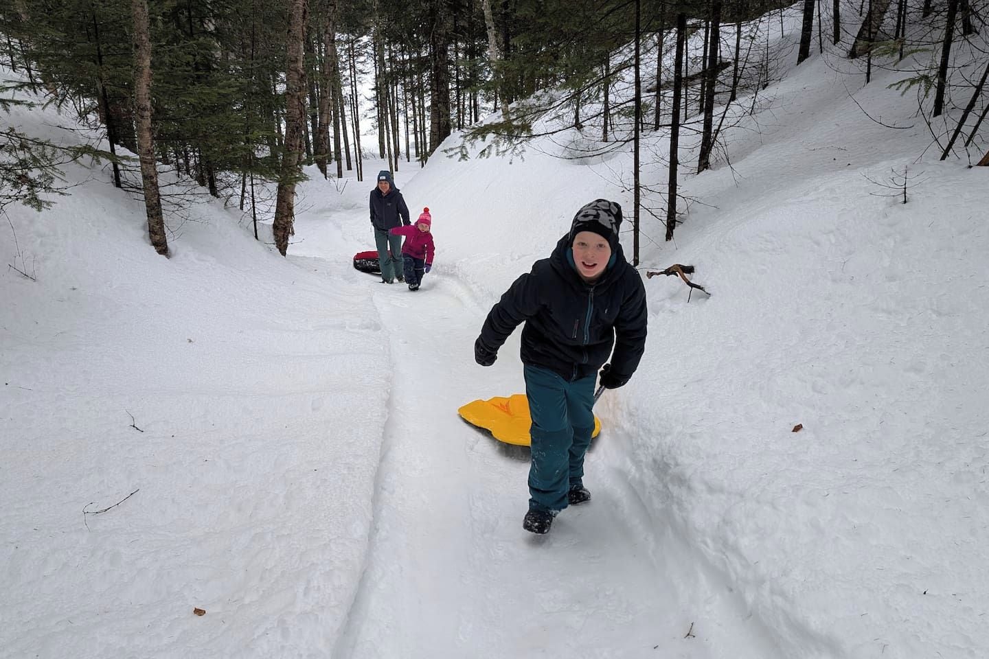 A snowy forested area with a person on a snowboard and another person in the background.