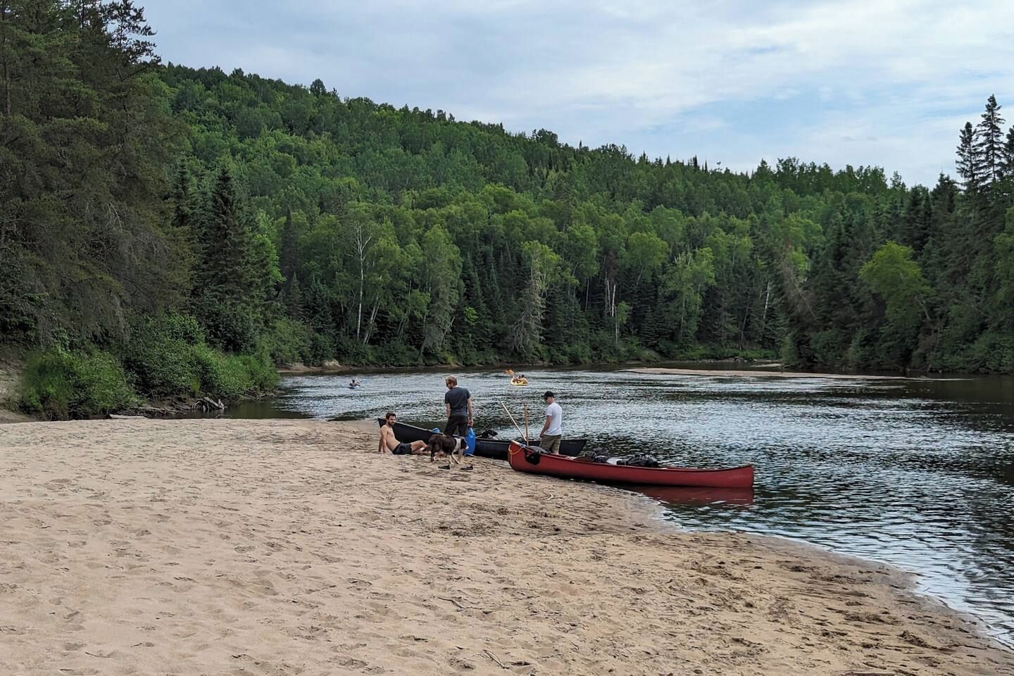 A sandy beach with a river, a red canoe, and people.