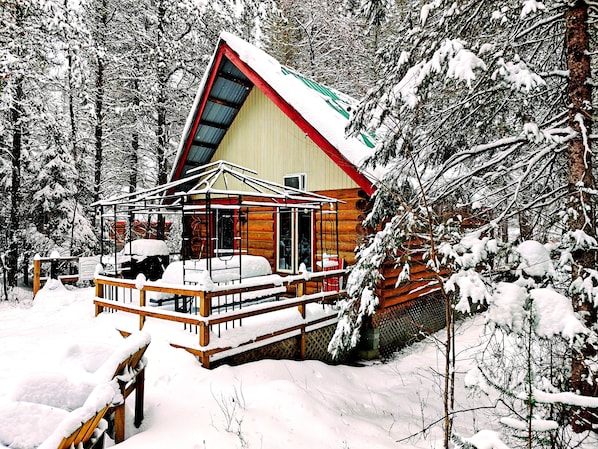 A log cabin with a balcony and a covered porch, surrounded by snow-covered trees.