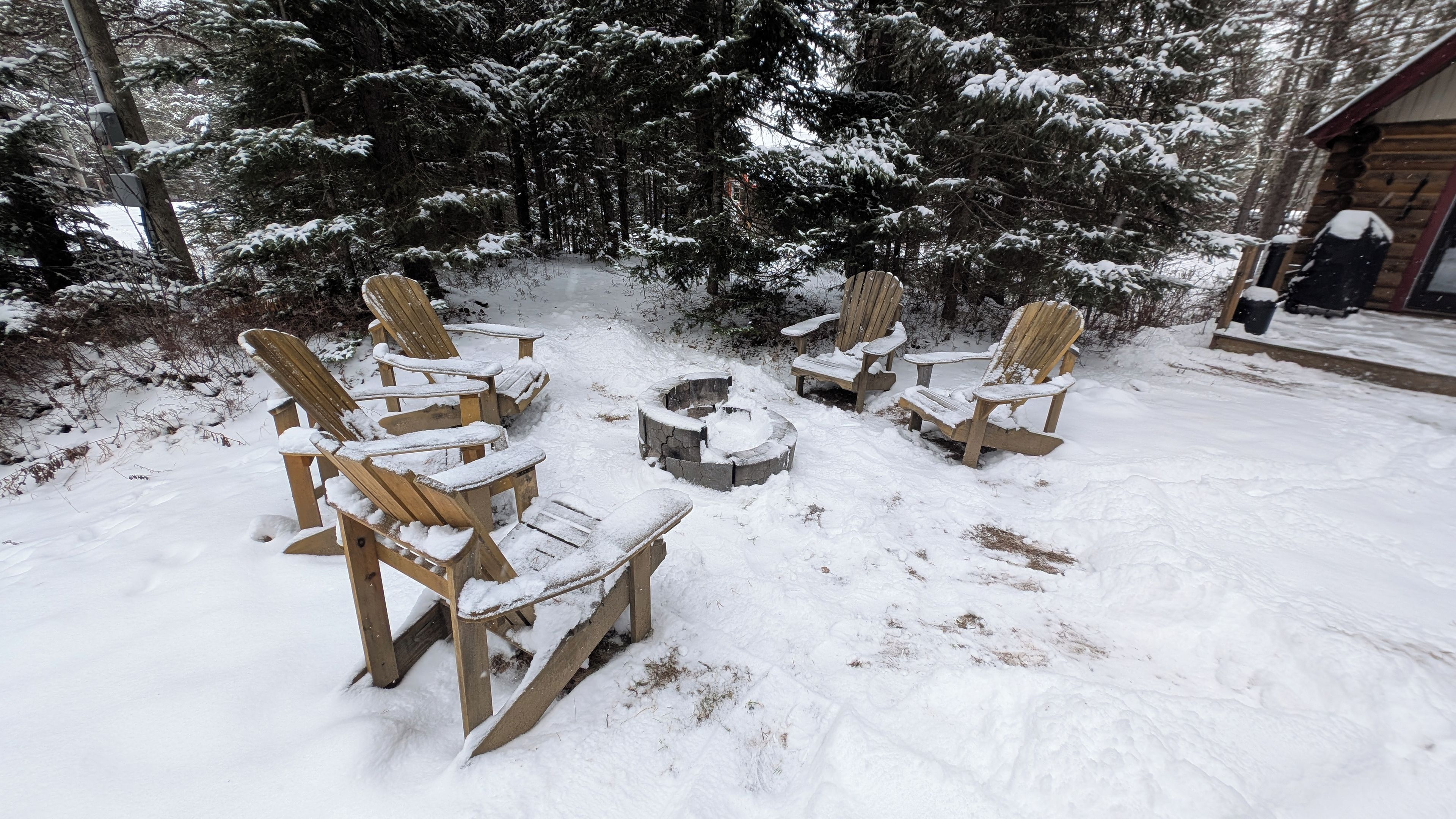 Un espacio al aire libre cubierto de nieve con cuatro sillas Adirondack de madera dispuestas alrededor de un hogar central.