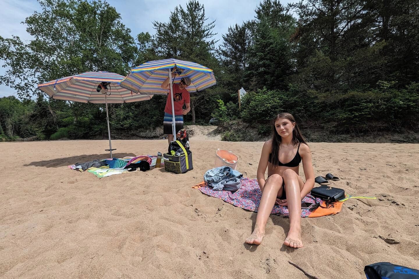 Escena de playa con una persona sentada sobre una toalla, dos sombrillas de playa a rayas y varios objetos de playa esparcidos.