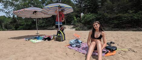 Escena de playa con una persona sentada sobre una toalla, dos sombrillas de playa a rayas y varios objetos de playa esparcidos.