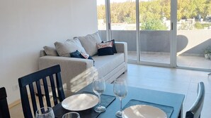 A dining area with a table set for two, a sofa in the background, and a view of trees and buildings through large windows. - Departamento Hermosa Vista Mendoza (Mendoza)