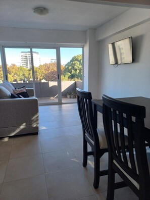 A modern living room with a sofa, a dining table, and chairs. There is a sliding glass door leading to a balcony with a view of trees and buildings. - Departamento Hermosa Vista Mendoza (Mendoza)