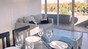 A modern dining area with a table set for four, a sofa with cushions, and a large sliding glass door leading to an outdoor balcony. - Departamento Hermosa Vista Mendoza (Mendoza)