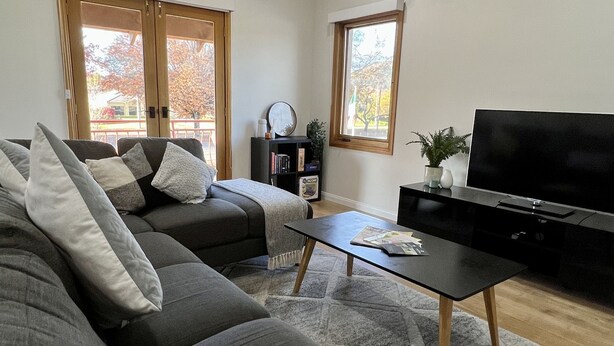 A modern living room with a grey sectional sofa, a black coffee table, and a flat-screen TV. There are two windows with curtains, a ceiling fan, and a small bookshelf. - Tuscan Villa in Myrtleford (Myrtleford)