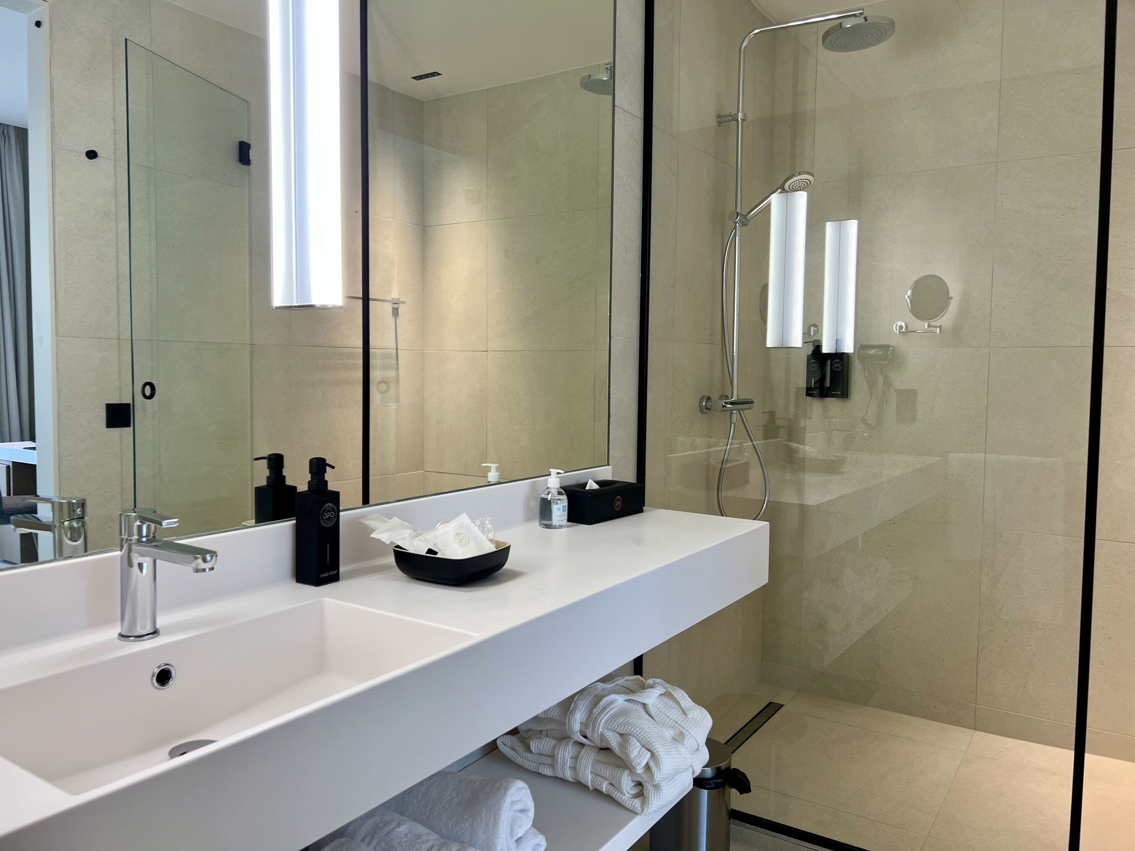 A modern bathroom with a large mirror, a white sink, a black soap dispenser, and a glass-enclosed shower area.